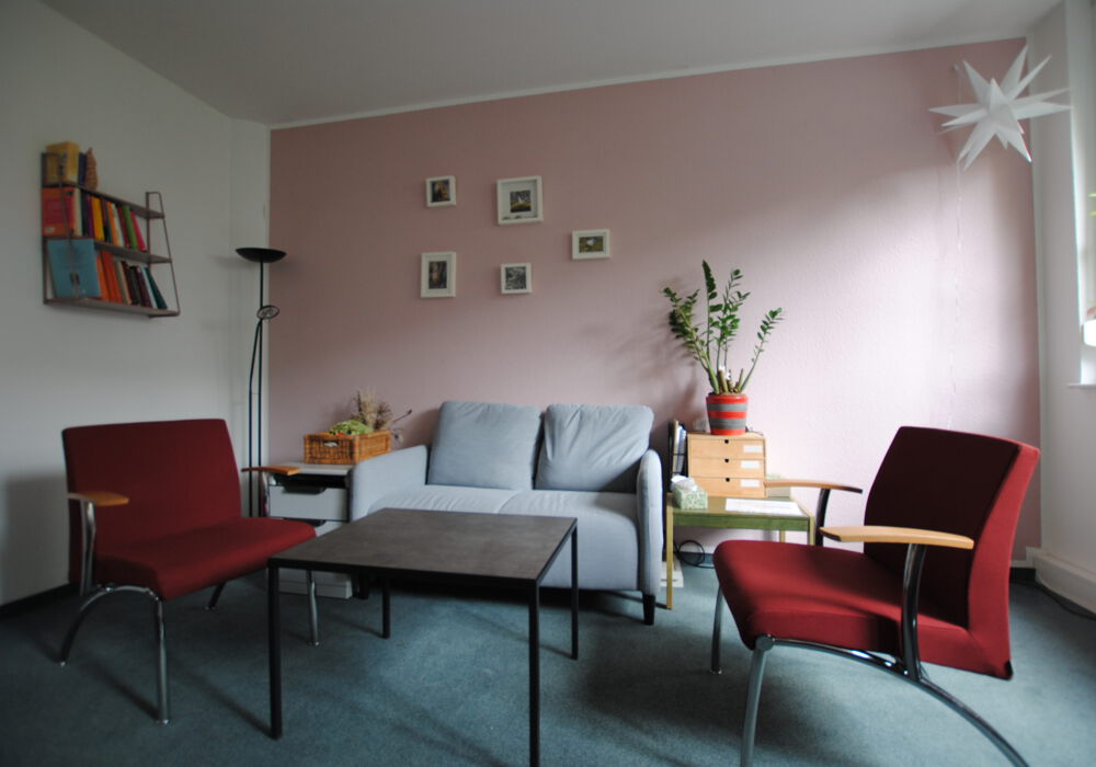 Beratungsraum des Teams der aufsuchenden Familientherapie AFT+ zwei rote Stühle, ein Tisch und ein graues Sofa stehen in einem gemütlich wirkenden Raum mit einer Pflanze und Bildern an den Wänden