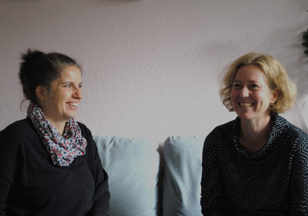 das Team der Aufsuchenden Familientherapie AFT+ zwei Kolleginnen aus dem Team der Aufsuchenden Familientherapie sitzen auf einem hellblauen Sofa und lächeln in die Kamera